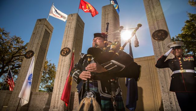 A bagpipe musician plays in front of the Armed Forces flags. A service man salutes behind him.