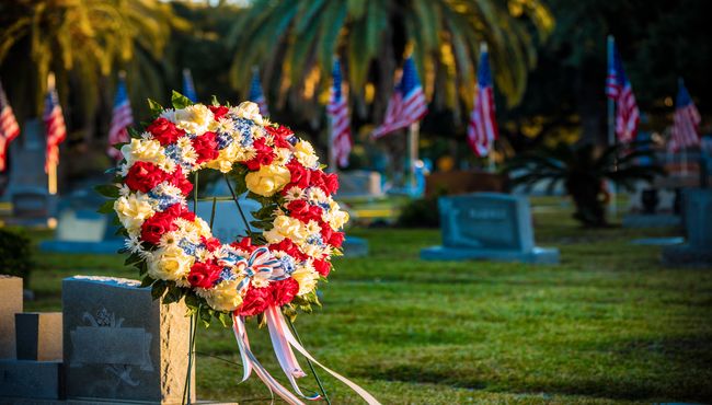 A patriotic floral wreath is displayed in a cemetery with upright markers and American flags