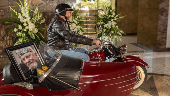 Motorcycle or biker riding into chapel for memorial service with sidecar containing portrait and urn. 