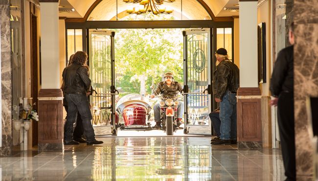 Motorcycle or biker riding into mausoleum chapel for memorial service with urn and portrait in sidecar and other bikers looking on.