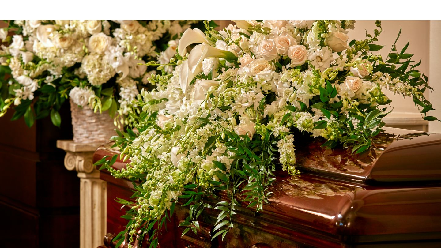 Spray of white roses and tulips laid on top of a wooden casket inside a funeral home. 