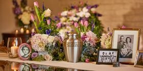 Display table with brass urn, pictures, flowers and glowing candle sitting on top of it at Pacific View